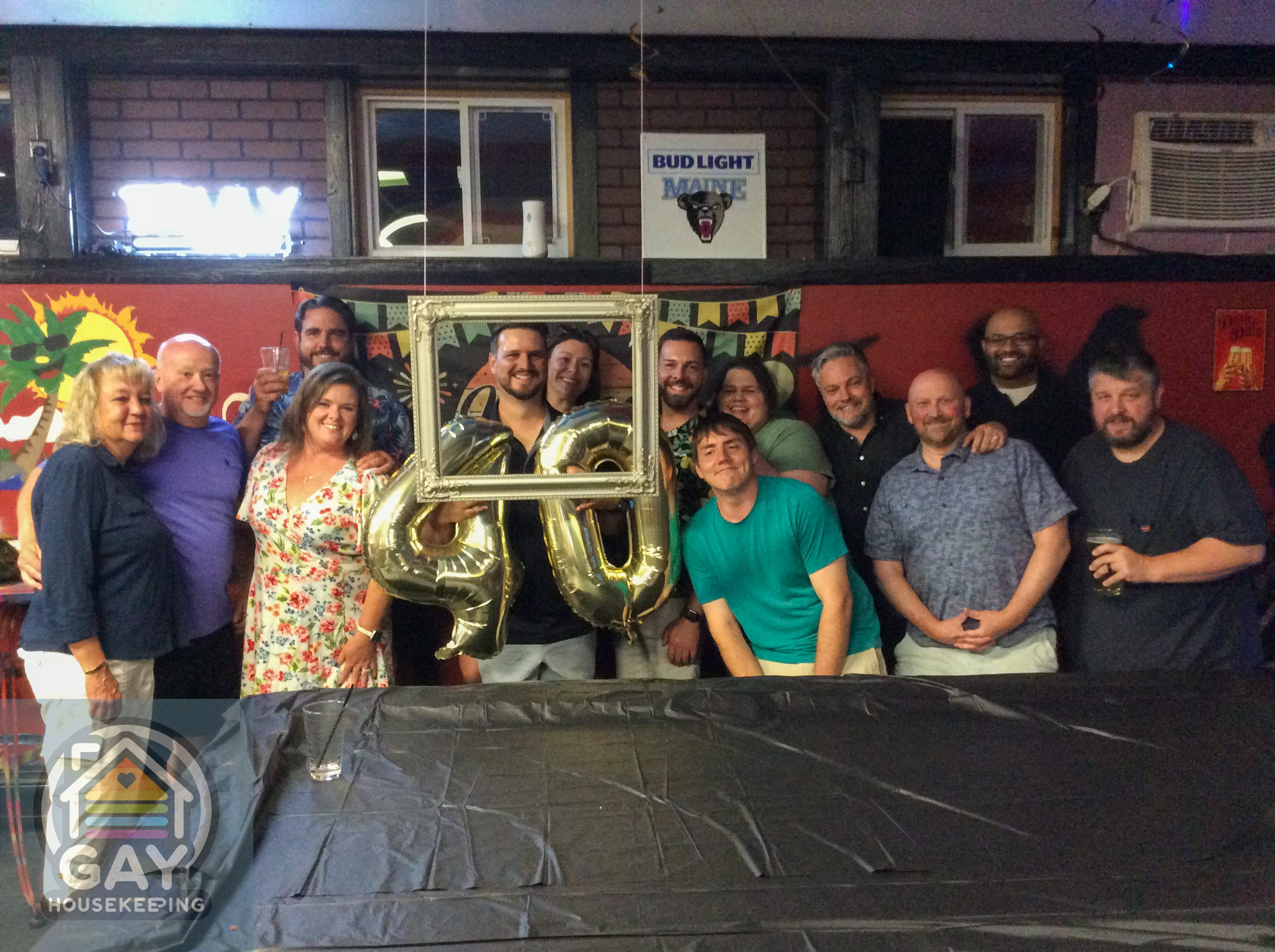 Friends crowded together at the birthday party, holding gold "40" balloons and a silver picture frame.