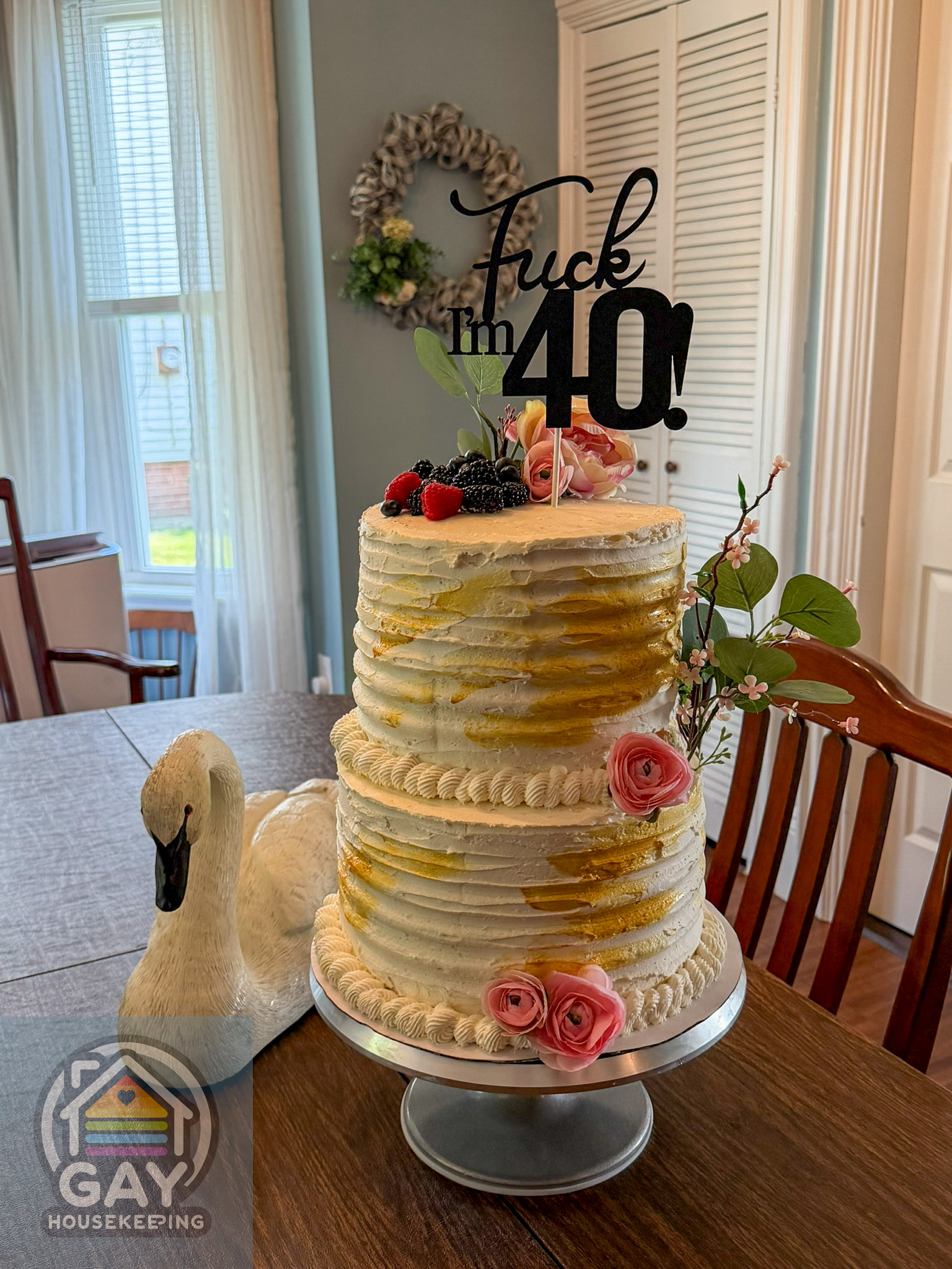 The finished two-tier cake with rustic gold-brushed ermine buttercream, pink flowers, fresh berries, and a black "Fuck, I'm 40!" topper.