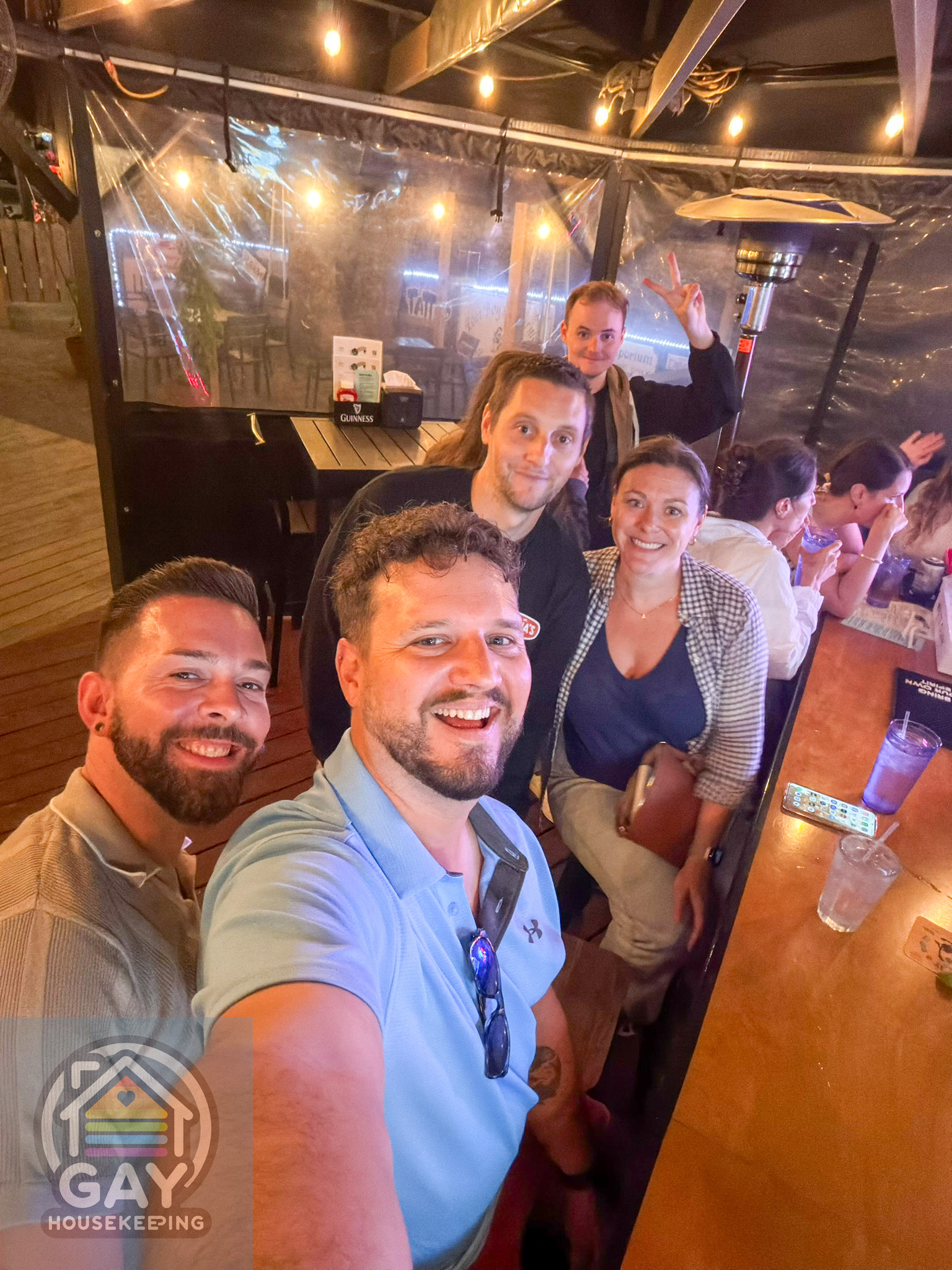 Selfie of four friends grinning at the bar under string lights, one throwing a peace sign in the back.