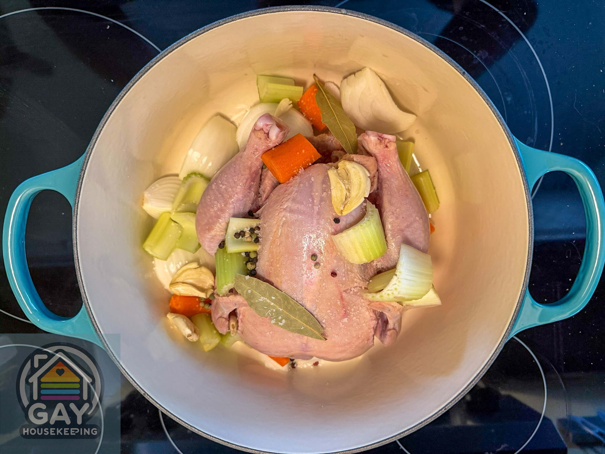 A whole raw chicken nestled in a teal Le Creuset Dutch oven with quartered onion, carrots, celery, garlic, bay leaves and peppercorns.