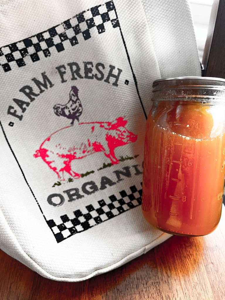 A mason jar filled with homemade stock is placed on a wooden table next to a kitchen towel with a 'Farm Fresh Organic' print featuring a pig and a chicken. The towel has a rustic checkerboard border. The amber hue of the stock indicates a rich, flavorful broth, and the jar is marked with measurements, suggesting the stock is freshly made and ready for use or storage.