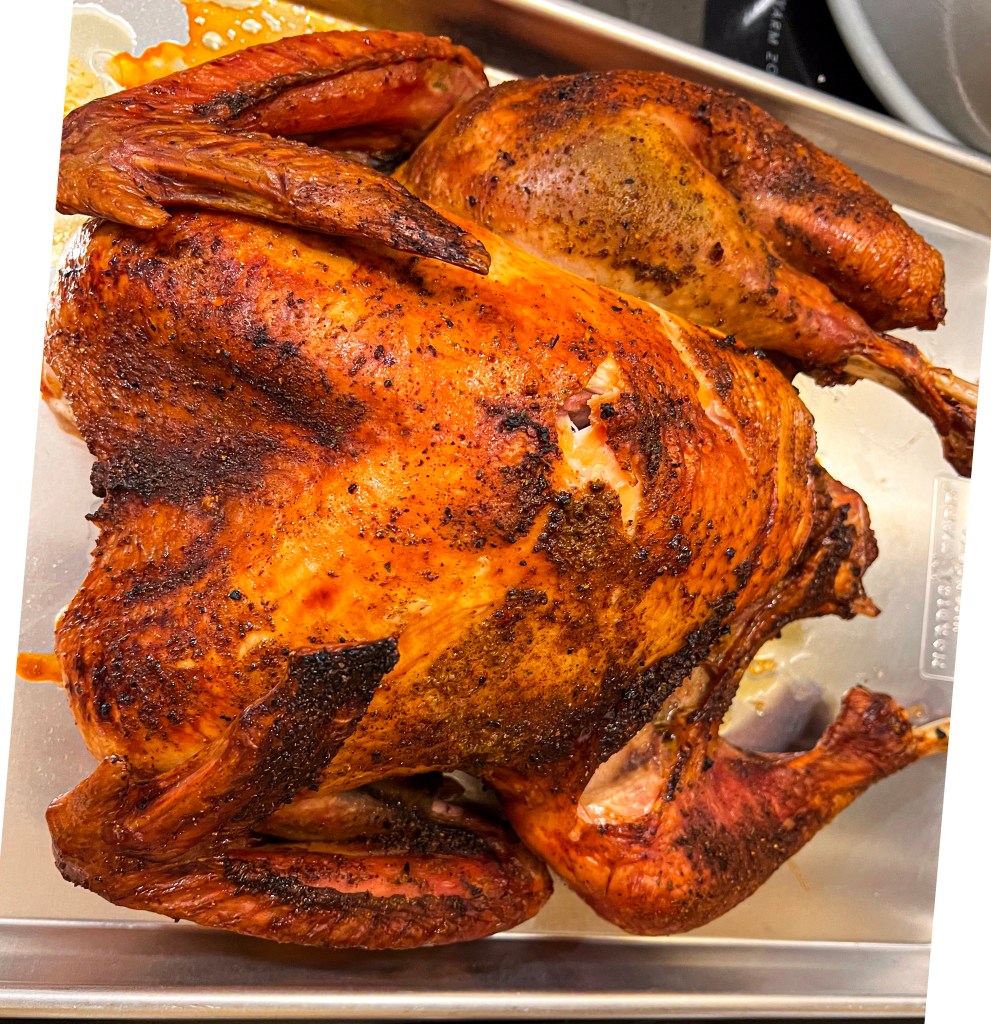 Golden-brown smoked turkey with crispy skin resting on a stainless steel tray, showcasing a well-cooked Thanksgiving feast centerpiece.