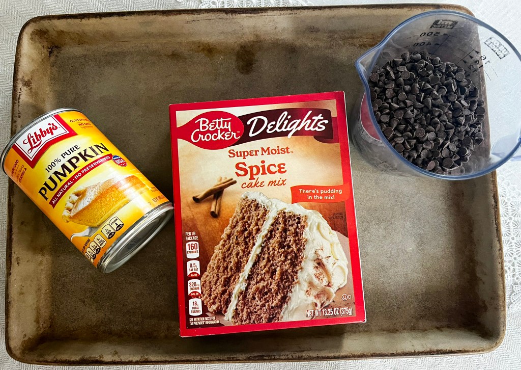Ingredients for pumpkin chocolate chip cookies laid out on a kitchen counter, including a can of pure pumpkin puree, a box of spice cake mix, and a measuring cup filled with chocolate chips next to a well-used baking sheet.