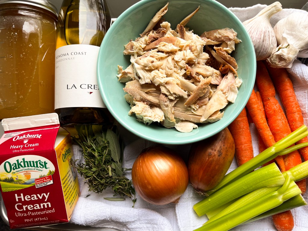 Ingredients for turkey soup including shredded turkey, carrots, celery, onion, garlic, fresh herbs, heavy cream, and chardonnay wine on a kitchen counter.