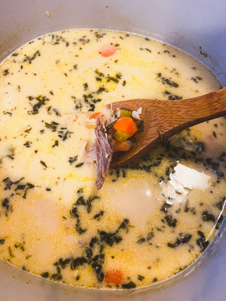 Creamy turkey soup with chunks of meat, vegetables, and herbs being stirred with a wooden spoon in a pot.