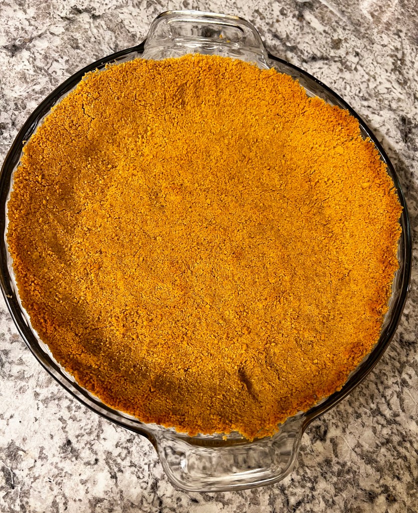 A freshly prepared graham cracker crust in a clear glass pie dish on a marble countertop.