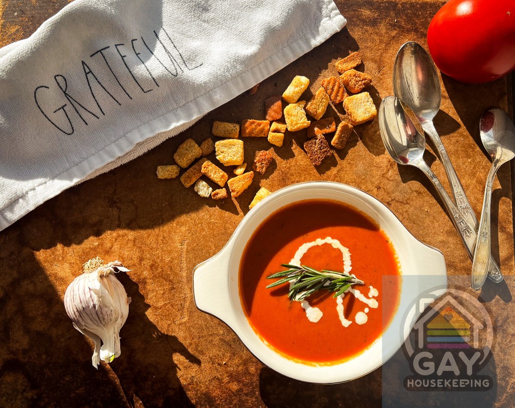 A bowl of creamy tomato soup garnished with a swirl of cream and fresh rosemary, accompanied by croutons, a whole tomato, garlic, and two spoons, set on a rustic surface with a napkin inscribed with 'GRATEFUL' and the logo of 'GAY Housekeeping'.