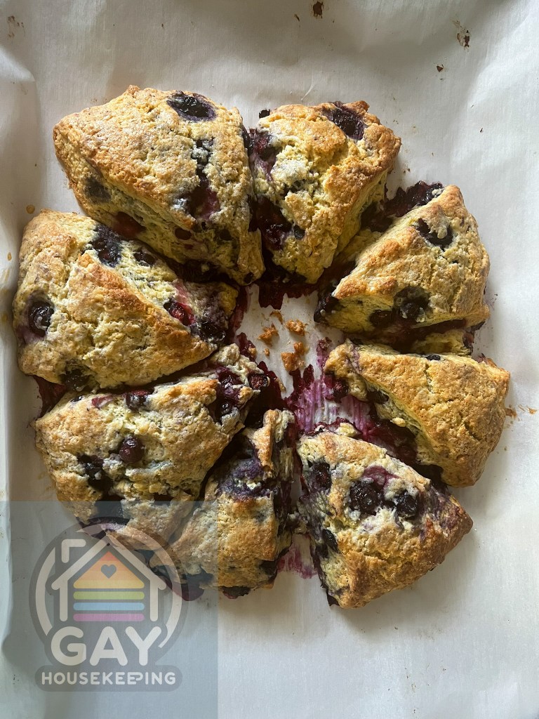 Freshly baked blueberry scones on parchment paper with a 'Gay Housekeeping' logo in the foreground.